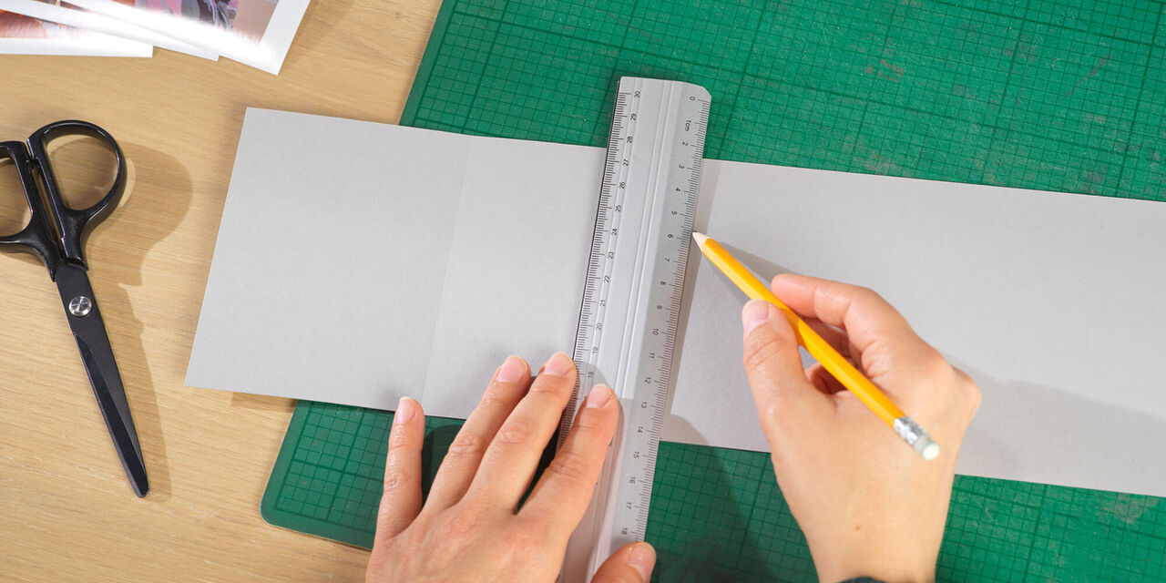 Dve roki s svinčnikom in ravnilom rišeta linijo za fotografijo na listu sivega kartona.