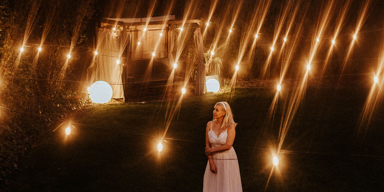 Ženska v beli obleki stoji pod nočnim nebom. Obdana je z ambientalno svetlobo, v ozadju pa je osvetljen paviljon.