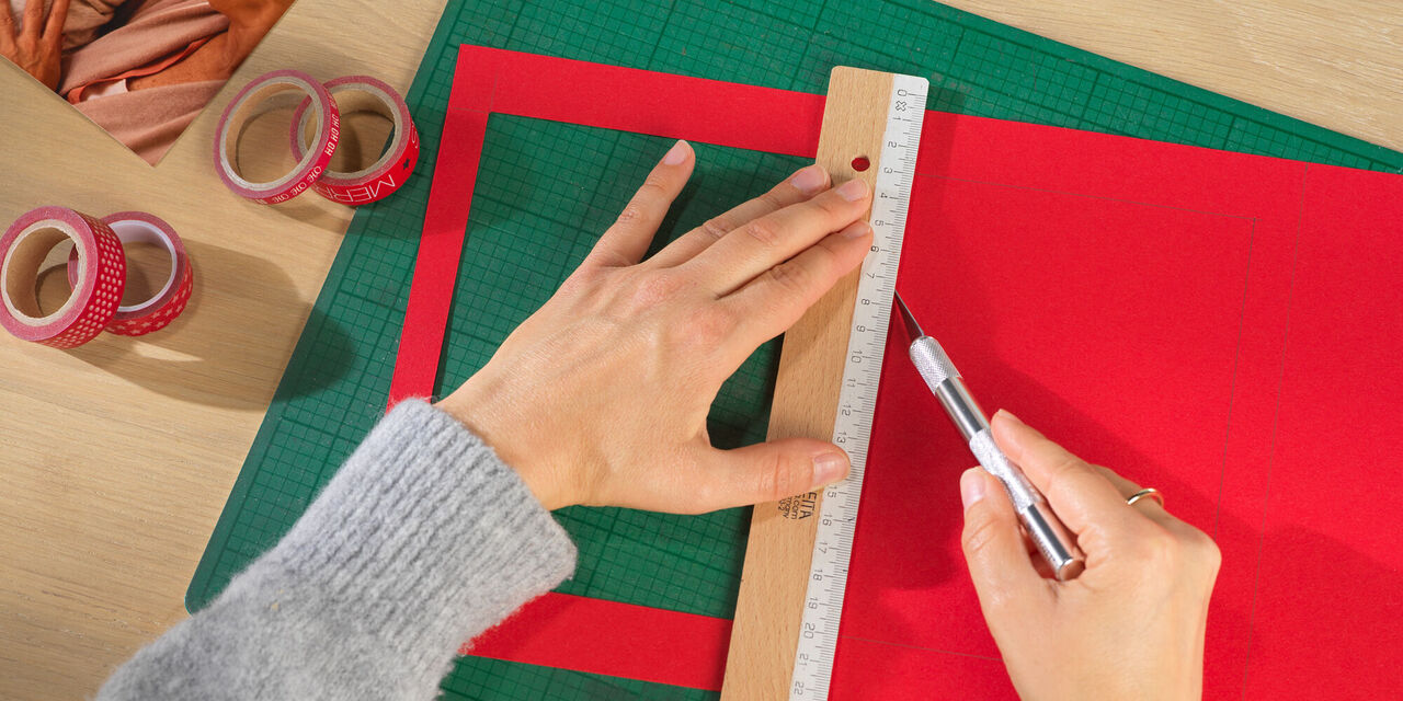 Dve roki držita ravnilo in svinčnik. Eden od štirih okvirjev, ki jih je treba izdelati, je že izrezan iz rdečega kartona.